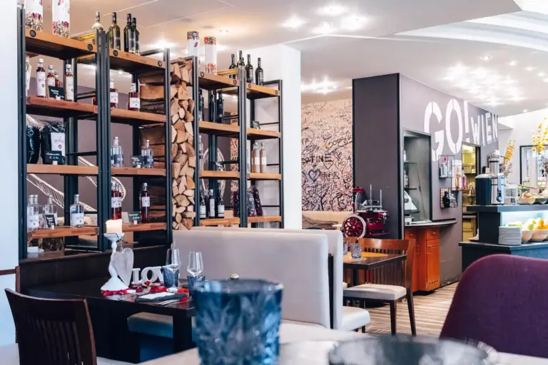 Ein modernes Restaurant mit Holzregalen, in denen Flaschen und Brennholz ausgestellt sind, gemütlichen Sitzgelegenheiten, gedeckten Tischen und einer Wand mit dekorativer Schrift. Die Atmosphäre ist hell und einladend.