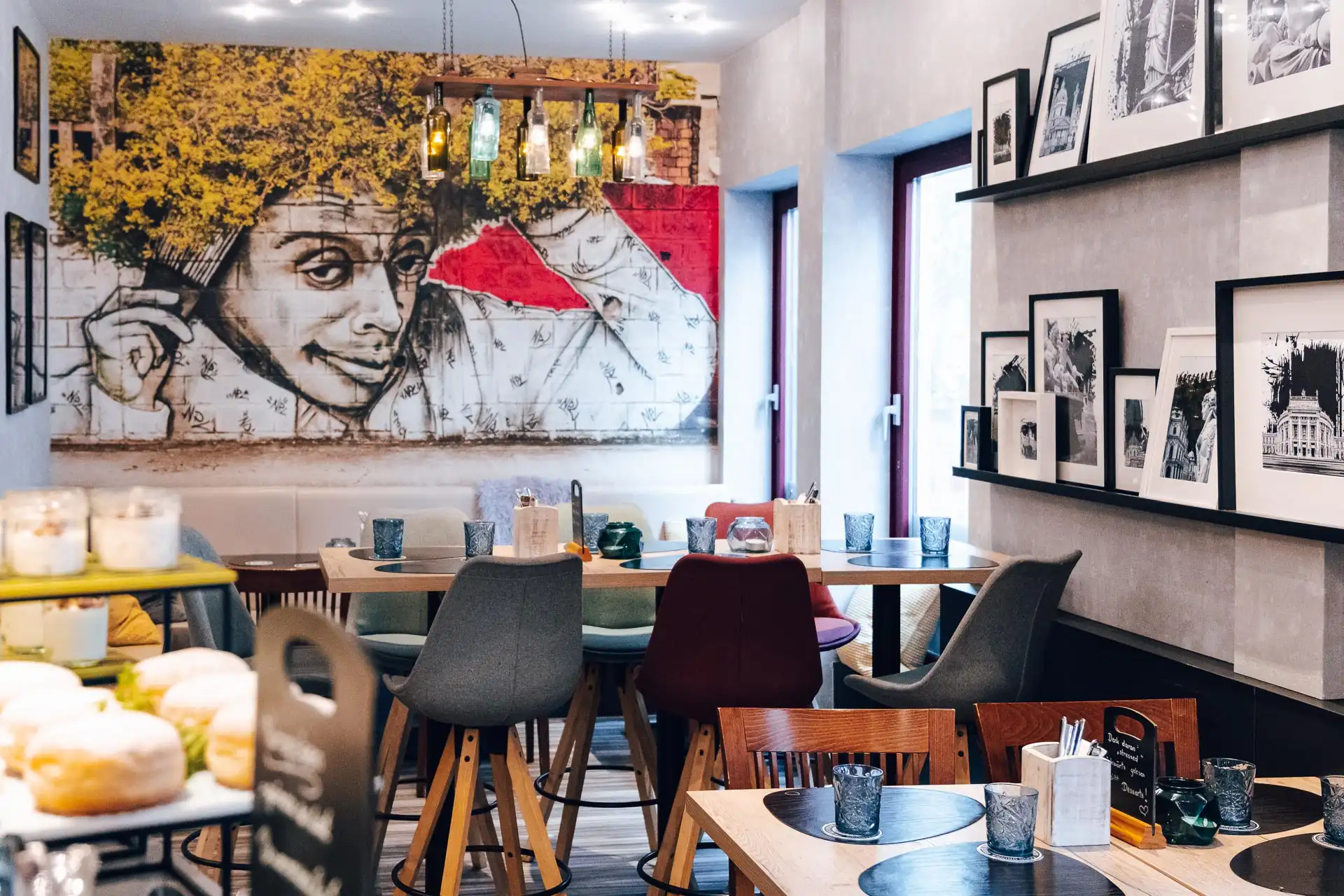 Ein gemütliches Café mit moderner Einrichtung, bunten Stühlen, runden Tischen, gerahmten Schwarz-Weiß-Fotos an der Wand und einem großen Wandgemälde mit einer Person, die ihre Hände im Haar hat, im Hintergrund.