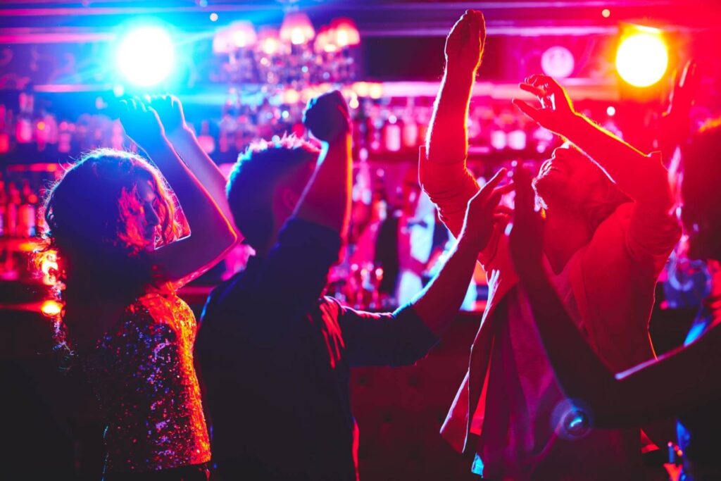 Energisch tanzende Menschen unter bunten Neonlichtern in einem lebhaften Nachtclub in Wien, perfekt zum Geburtstag feiern, mit einer Bar und Flaschen im Hintergrund.