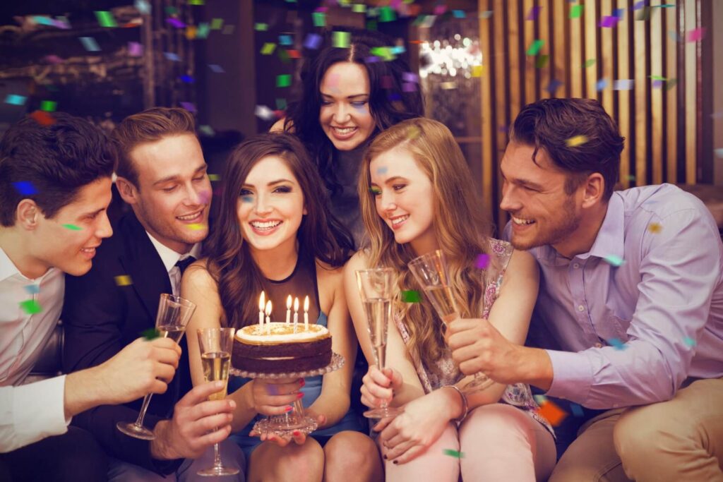 Eine Gruppe von sechs jungen Erwachsenen feiert drinnen in einer trendigen Geburtstagslocation in Wien einen Geburtstag. Die Frau in der Mitte lächelt über eine Torte mit brennenden Kerzen, während andere Sektgläser halten und Konfetti um sie herum fällt. Alle sehen fröhlich und festlich aus.