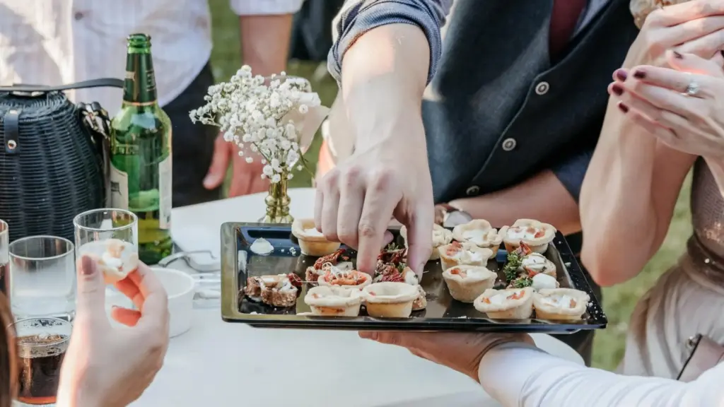 Eine Gruppe von Gästen wählt Häppchen von einem Tablett aus, das mit verschiedenen Fingerfoods gefüllt ist - die perfekte Inspiration, wenn Sie eine Geburtstagsparty planen. Eine kleine Vase mit weißen Blumen, Getränke und eine schwarze Tasche stehen auf dem Tisch.