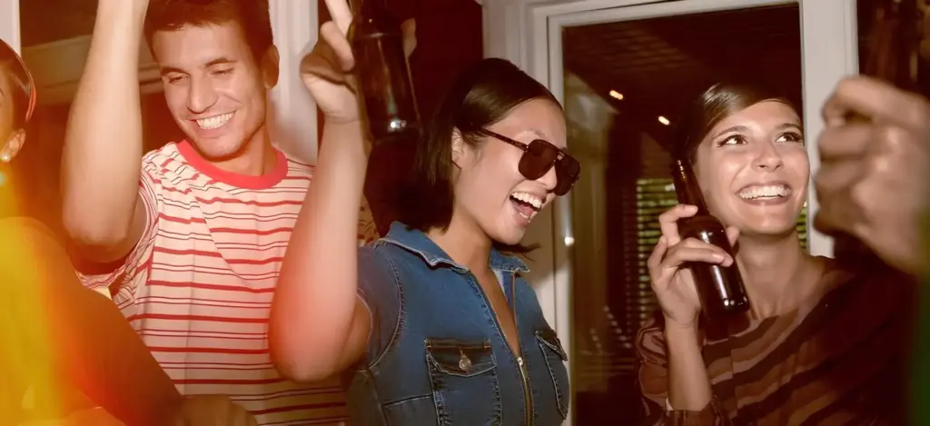 Drei junge Erwachsene lächeln und tanzen auf einer Party, in der Hand Getränke. Eine Frau trägt eine Sonnenbrille und ein Jeansoberteil, die andere ein gestreiftes Hemd - die perfekte Inspiration, wenn Sie eine Geburtstagsparty mit warmer Beleuchtung und lebendiger Stimmung planen möchten.