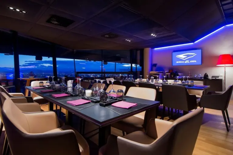 Generali Arena Ein moderner Restaurantbereich mit ordentlich gedeckten Tischen, beigen Stühlen, großen Fenstern, die eine Stadtlandschaft in der Abenddämmerung zeigen, und einem beleuchteten Schild an der Wand mit der Aufschrift "GENERAL APS ARENA".