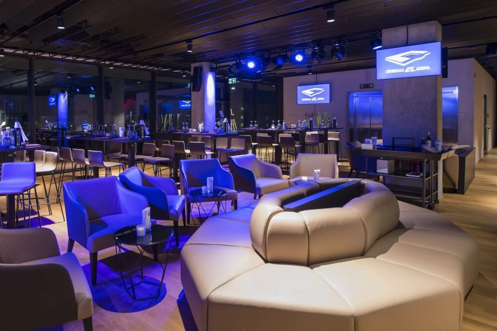 Moderner Lounge-Bereich mit runden beigen Sofas, kleinen Tischen und gedämpfter blauer Beleuchtung. Im Hintergrund sind große Fenster, Hochtische und ein Bildschirm mit dem Logo der Generali-Arena zu sehen.