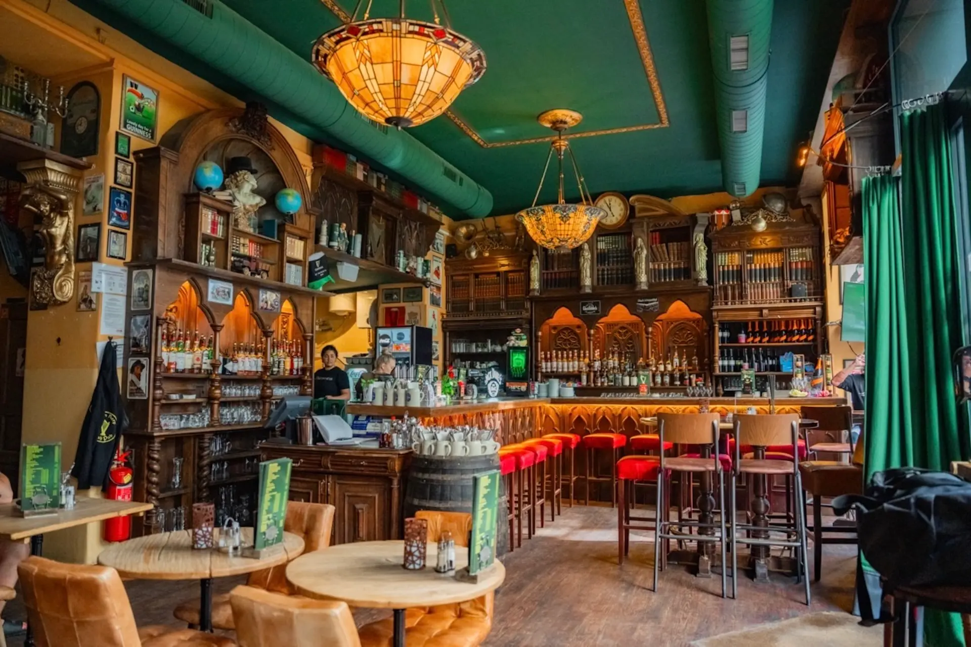 Eine gemütliche Bar mit verschnörkelten Holzarbeiten, roten Barhockern, runden Holztischen und Kronleuchtern. Gläser und Flaschen stehen in den Regalen hinter der Bar. Eine Person steht hinter dem Tresen. Grüne Vorhänge und Dekoartikel bringen Farbe ins Spiel.
