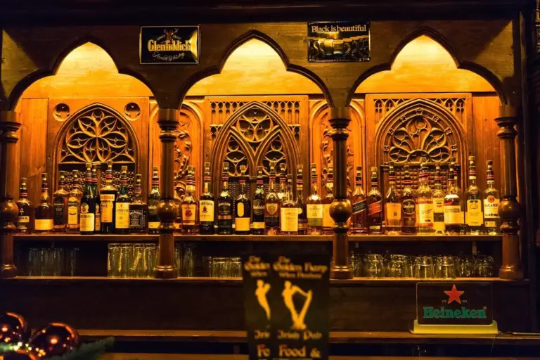 Elegante Holzbar mit verschnörkelten Bögen und Regalen mit verschiedenen Schnapsflaschen und Glaswaren. Die warme gelbe Beleuchtung unterstreicht die gotischen Holzarbeiten und schafft eine gemütliche Atmosphäre. Schilder für Glenfiddich und Heineken sind sichtbar.