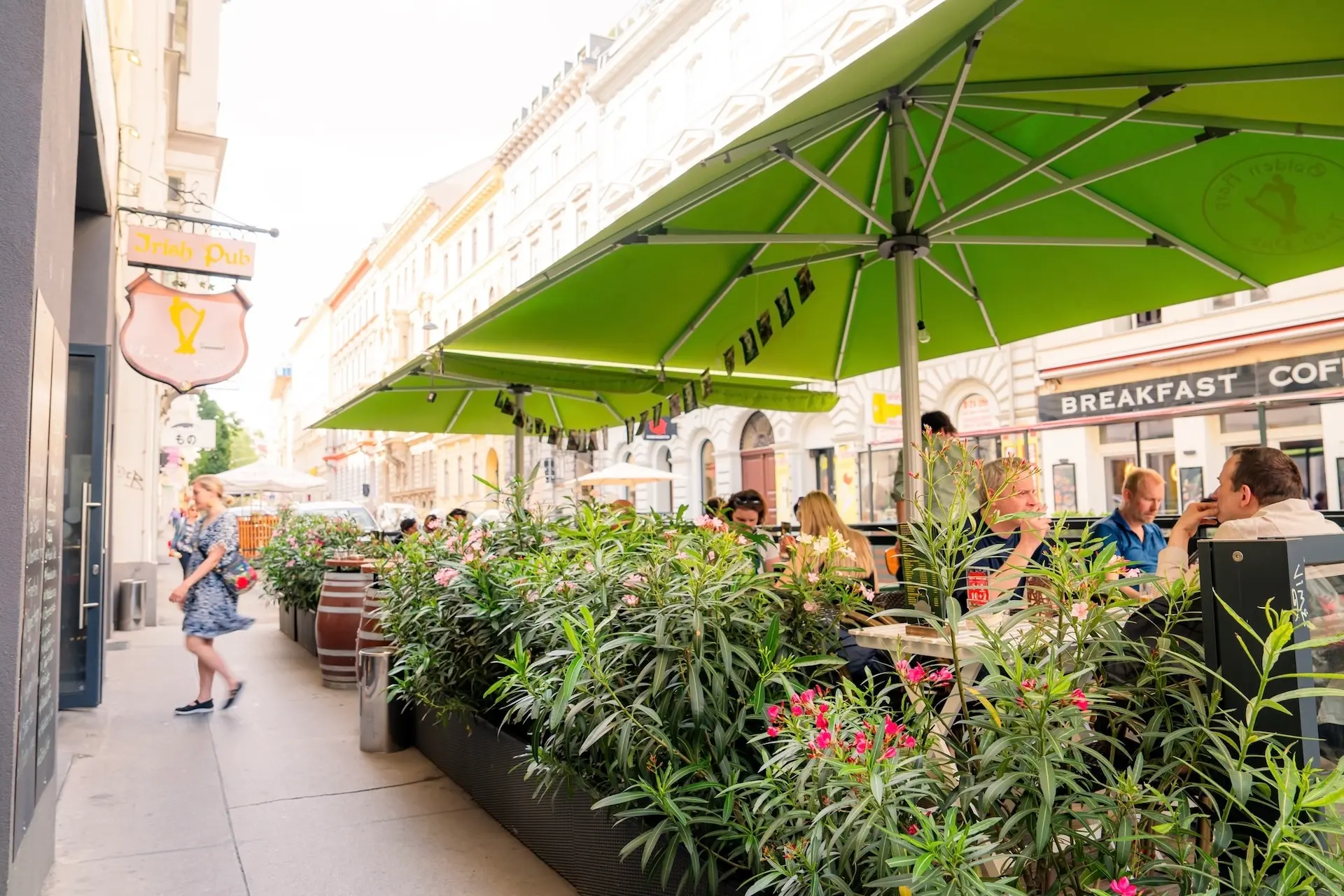 Menschen, die an Cafétischen im Freien unter großen grünen Sonnenschirmen in einer belebten Straße essen, umgeben von Topfpflanzen, mit einer vorbeigehenden Person und den Schildern eines nahe gelegenen Geschäfts.