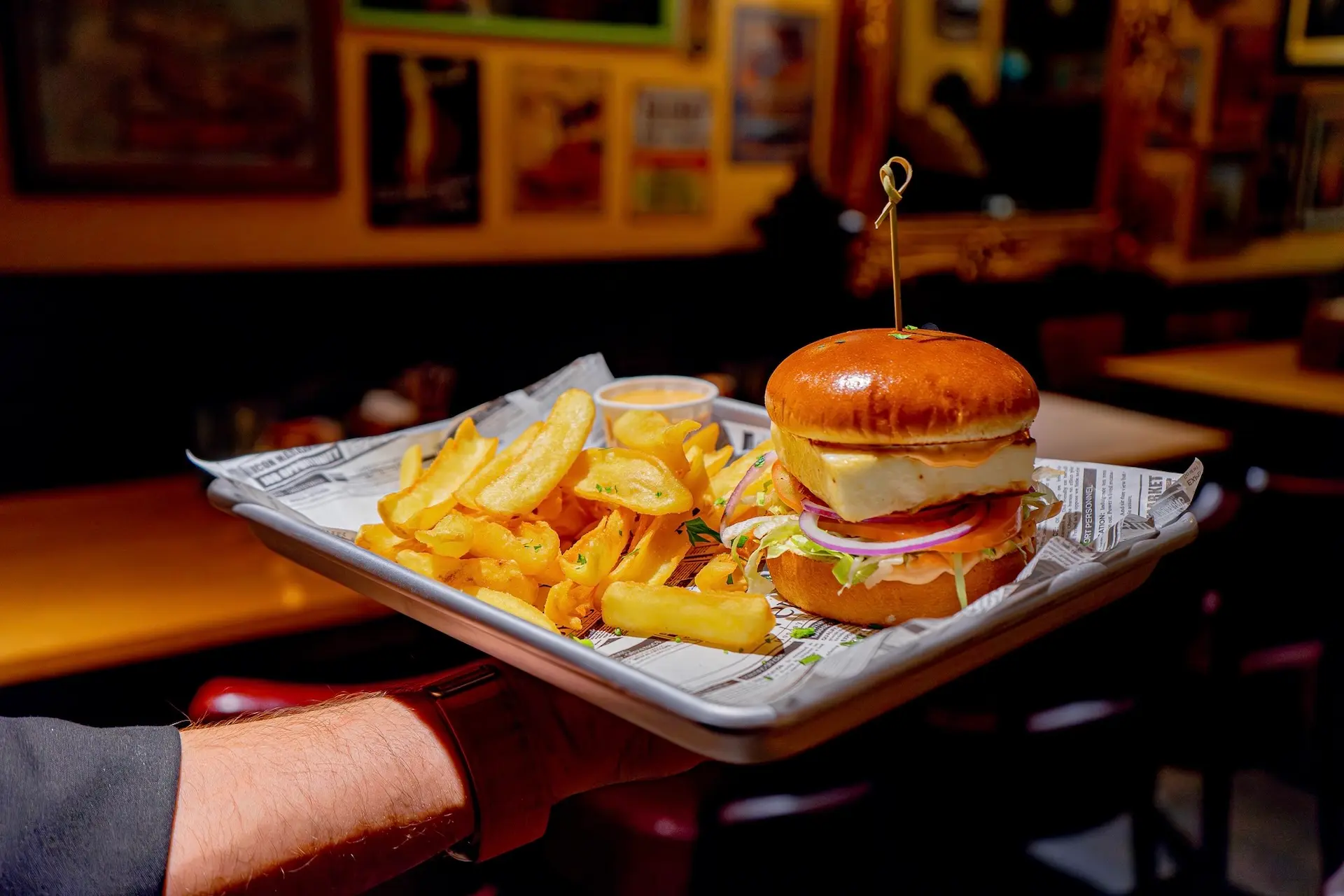 Eine Hand hält ein Tablett mit einem Burger, der mit Käse, Salat, Tomaten und Zwiebeln belegt ist, zusammen mit dick geschnittenen Pommes frites und einem kleinen Becher mit Dip-Soße, serviert auf Papier in einem gemütlichen, warm beleuchteten Restaurant.