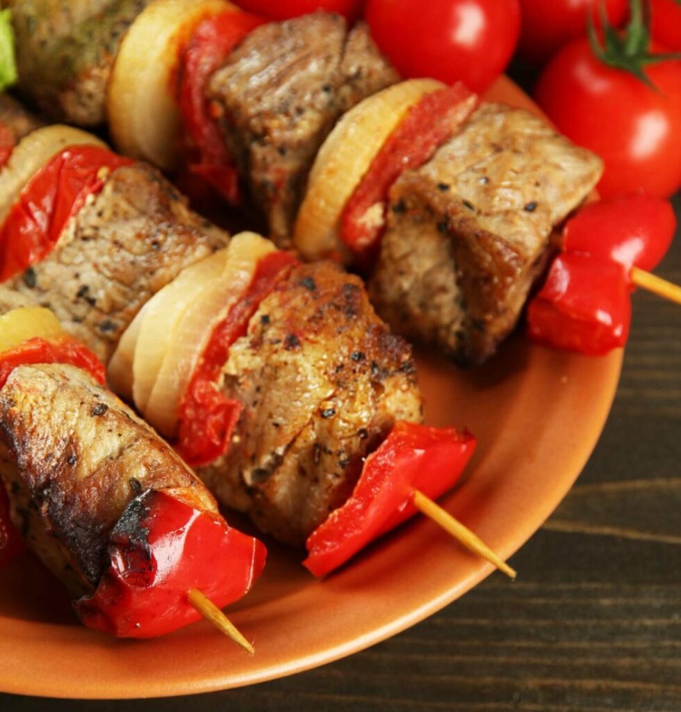 Grillen bringt die reichen Aromen von gegrilltem Fleisch, roten Paprikastücken und Zwiebeln zur Geltung, die auf Holzspieße aufgespießt und auf einem braunen Teller serviert werden, mit frischen Tomaten im Hintergrund.