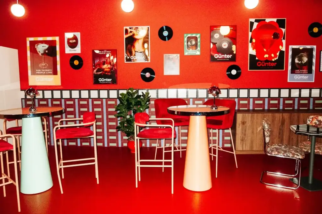 Ein Cafe im Retrostil mit roten Wänden, Tischen und Stühlen. Schallplatten und Poster schmücken die Wand, und zwischen den Tischen steht eine Grünpflanze. Hängelampen werfen ein warmes Licht auf die lebendige, eklektische Einrichtung.