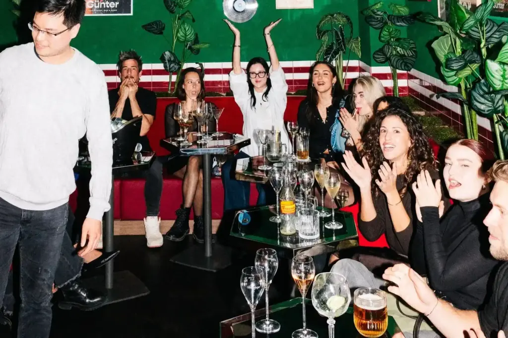 Eine Gruppe von Menschen sitzt an Tischen mit Getränken, lächelt, klatscht und jubelt in einer lebhaften, bunten Bar mit grünen Wänden und Topfpflanzen. Eine Person steht auf der linken Seite, teilweise außerhalb des Bildes.