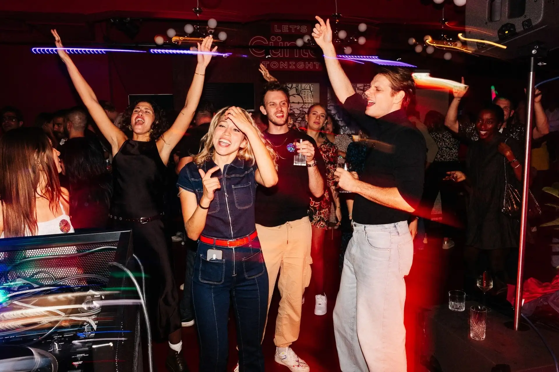 Eine lebhafte Gruppe von Menschen singt und tanzt enthusiastisch in einer Karaoke-Bar, mit bunten Lichtern und Getränken in der Hand, die die energiegeladene Atmosphäre eines unterhaltsamen Abends einfangen.