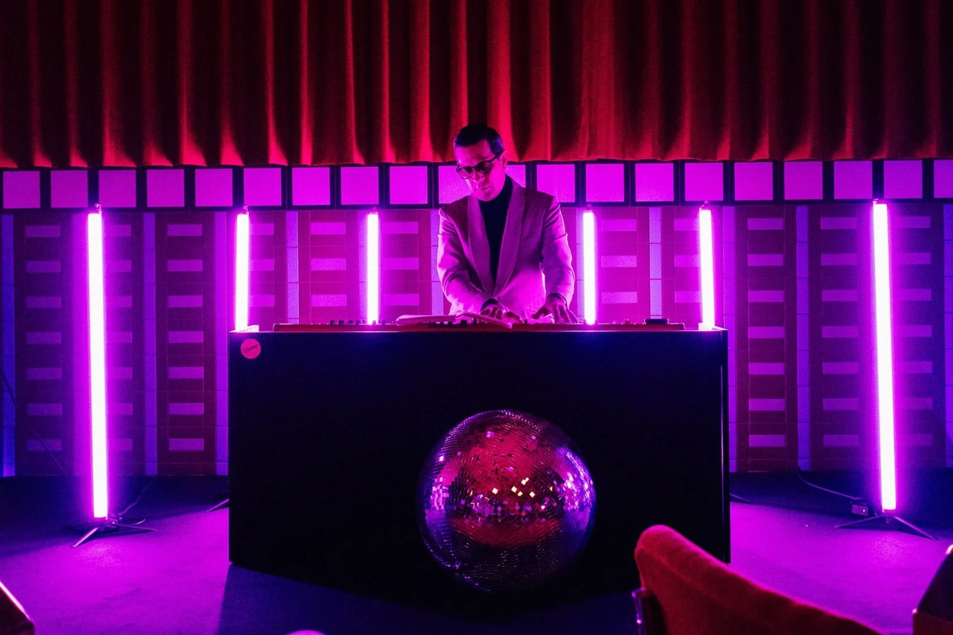 Ein DJ spielt Musik hinter einer großen Kabine mit einer spiegelnden Discokugel davor, die von hellvioletten vertikalen Lichtern beleuchtet wird. Der Hintergrund besteht aus gemusterten Wänden und roten Vorhängen, die eine lebhafte Partyatmosphäre schaffen.