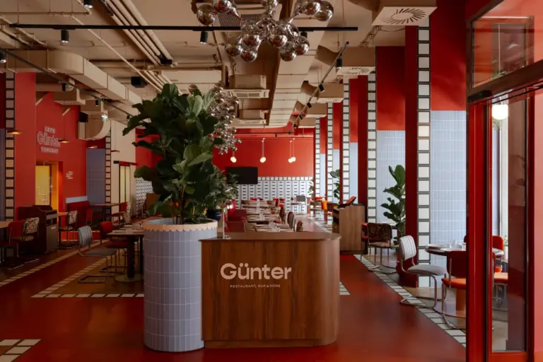 Modernes Restaurantinterieur mit roten Böden und Wänden, geometrischen Kachelakzenten und großen Grünpflanzen. Am Eingang befindet sich ein hölzerner Empfangstresen mit der Aufschrift "Günter". Tische und Stühle sind ordentlich im Raum verteilt.