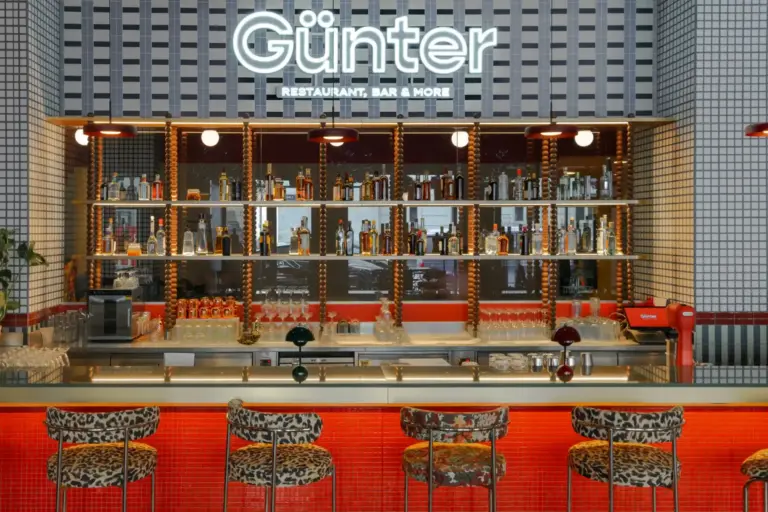 Eine moderne Bar mit gemusterten Hockern, einem roten Tresen, Regalen mit verschiedenen Schnapsflaschen, hängenden Gläsern und einer Leuchtreklame mit der Aufschrift Günter Restaurant, Bar & More an einer geometrisch gekachelten Wand.