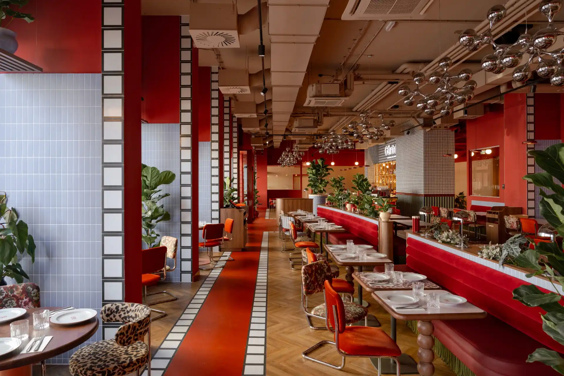 Ein modernes Restaurant mit roten Wänden, roten und orangefarbenen Stühlen, weiß gekachelten Säulen und hängenden Metallleuchten. Die Tische sind zum Essen gedeckt, und im ganzen Raum sind Pflanzen aufgestellt.