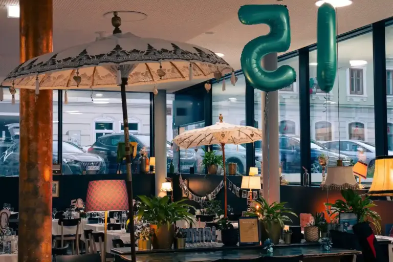 Habibi & Hawara, ein gemütlich eingerichtetes Café mit Vintage-Lampen, Topfpflanzen, weißen Sonnenschirmen und großen, blaugrünen Luftballons in Form der Zahl 15. Durch die großen Fenster sieht man draußen geparkte Autos und Gebäude.
