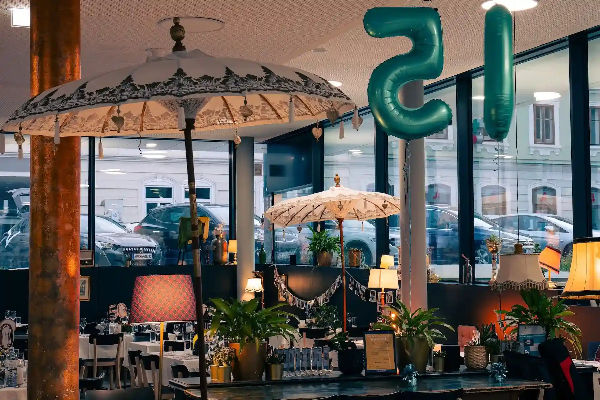 Habibi & Hawara, ein gemütlich eingerichtetes Café mit Vintage-Lampen, Topfpflanzen, weißen Sonnenschirmen und großen, blaugrünen Luftballons in Form der Zahl 15. Durch die großen Fenster sieht man draußen geparkte Autos und Gebäude.
