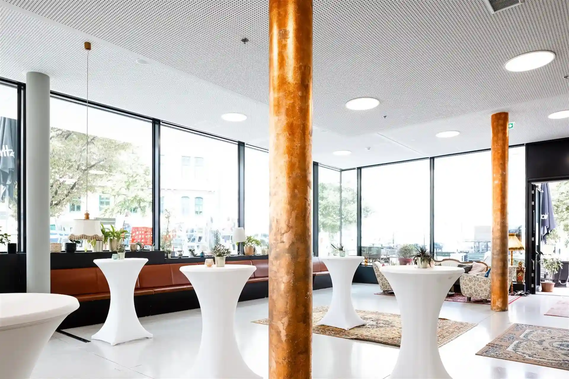 Eine helle, moderne Lounge mit großen Fenstern, runden, mit weißen Tüchern bedeckten Cocktailtischen, braunen, gepolsterten Sitzen an der Wand, orangefarbenen Säulen, Topfpflanzen und dekorativen Teppichen auf weißem Boden.