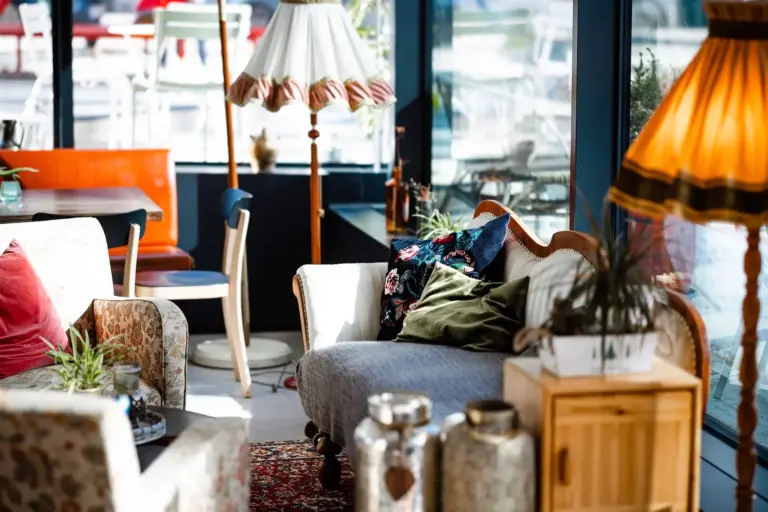 Ein gemütlicher, sonnendurchfluteter Raum mit Vintage-Möbeln, gemusterten Sesseln, einem Holzschrank, großen Lampen, Pflanzen und dekorativen Kissen auf einem Sofa in der Nähe großer Fenster.
