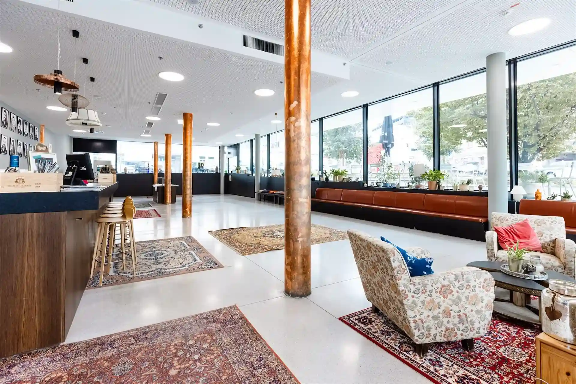 Eine geräumige, moderne Hotellobby mit großen Fenstern, Holzsäulen, gemusterten Teppichen, einer langen braunen Lederbank und einer Sitzecke mit geblümten Sesseln und einem kleinen Tisch in einer hellen, einladenden Umgebung.