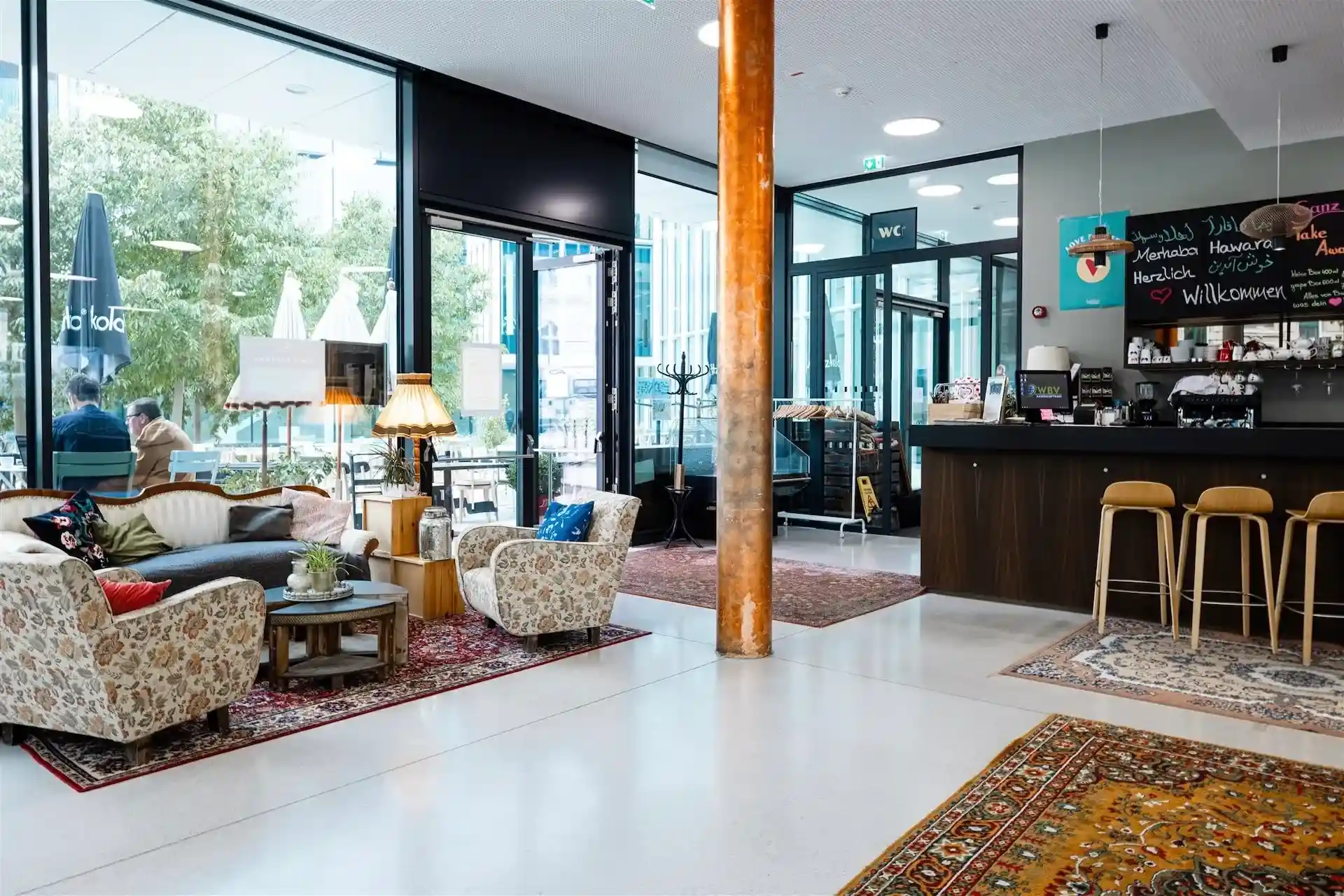 Ein gemütliches Café mit gemusterten Sesseln, Sofas und Teppichen, warmer Beleuchtung, einem Bartresen mit Hockern und großen Fenstern, die den Blick auf einen Sitzbereich im Freien und Bäume freigeben.