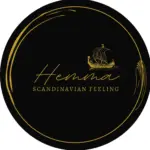 Schwarzes, kreisförmiges Logo mit goldenen Akzenten, mit einer Abbildung eines Wikingerschiffs über dem Wort hemma in Schreibschrift und SCANDINAVIAN FEELING in Großbuchstaben darunter. Elegante goldene geschwungene Linien verzieren die Ränder.