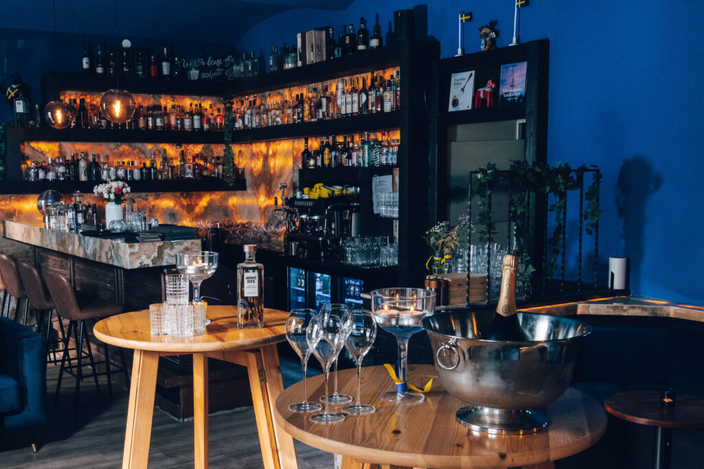 Eine stilvolle Bar mit Regalen voller Schnapsflaschen, einem Marmortresen, Hängelampen und einem runden Holztisch mit Weingläsern, einer Champagnerflasche in einem Eiskübel und Kerzen vor einer blauen Wand.