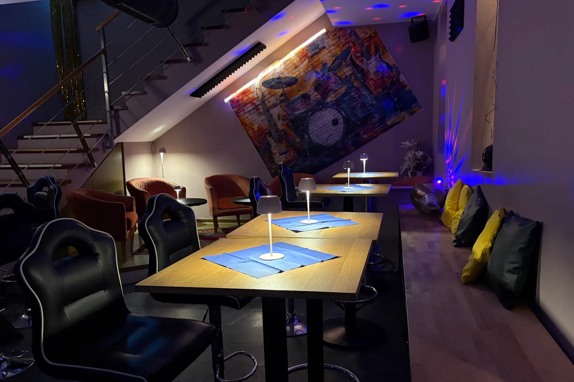 Eine gemütliche, schwach beleuchtete Lounge mit modernen Möbeln, kleinen Tischen mit blauen Tischsets, gepolsterten Sesseln, Wandbildern eines Schlagzeugs und bunten Kissen, die an der Wand unter einer Treppe angebracht sind.