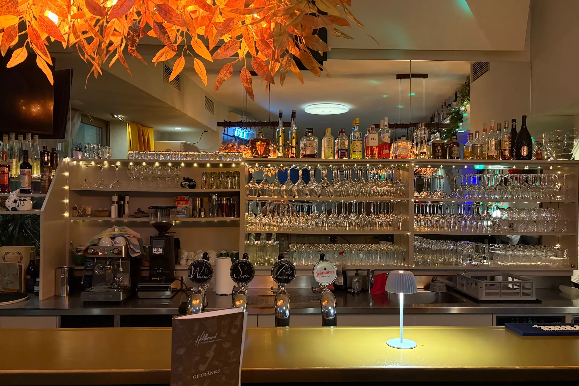 Ein gut beleuchteter Bartresen mit verschiedenen Gläsern, Alkoholflaschen und einer Kaffeemaschine dahinter. Warme Beleuchtung, Herbstlaubdekoration und Lichterketten schaffen eine gemütliche, einladende Atmosphäre.