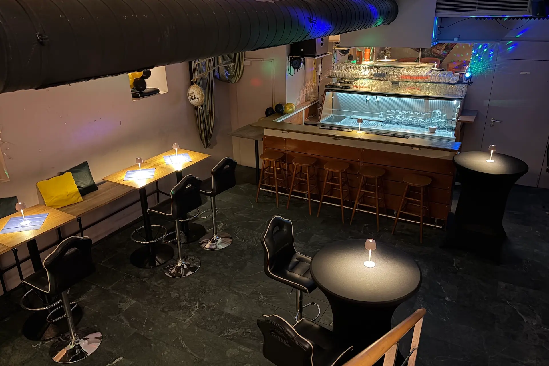 Eine moderne Bar mit hohen Tischen und Stühlen, gedämpfter Beleuchtung und einem langen Tresen mit Barhockern. Auf jedem Tisch stehen kleine Lampen, und an den Wänden spiegeln sich bunte Lichter.