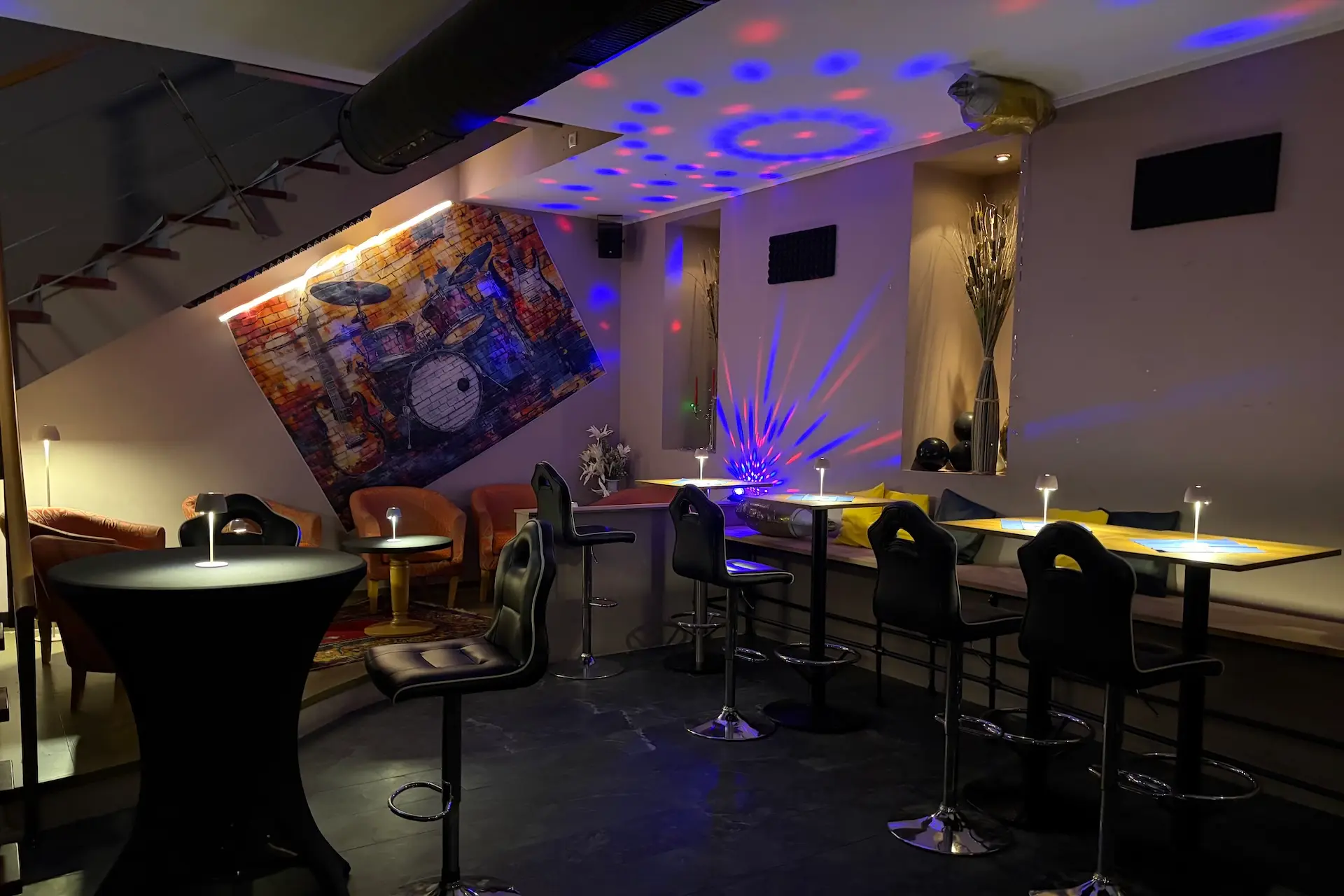 Eine moderne Bar mit hohen Tischen und Stühlen, bunten Scheinwerfern an der Decke, einem großen Gemälde mit Trommeln an der Wand und einer gemütlichen, gedämpften Atmosphäre.