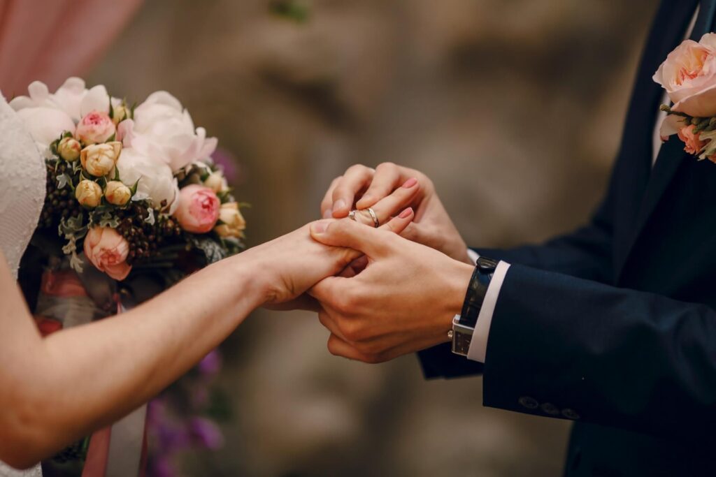 Nahaufnahme einer Braut und eines Bräutigams, die während einer Hochzeitszeremonie in einer beeindruckenden Hochzeitslocation in Wien ihre Ringe austauschen. Die Braut hält einen Strauß rosa und weißer Blumen, während der Bräutigam einen Anzug mit einer Boutonnière trägt.