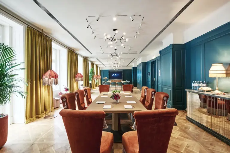 Eleganter Besprechungsraum mit einem langen Tisch, umgeben von rostfarbenen Plüschstühlen, goldenen Vorhängen, grünen Wänden, eklektischen Lampen, Zimmerpflanzen und einem großen Bildschirm am anderen Ende des Raums.