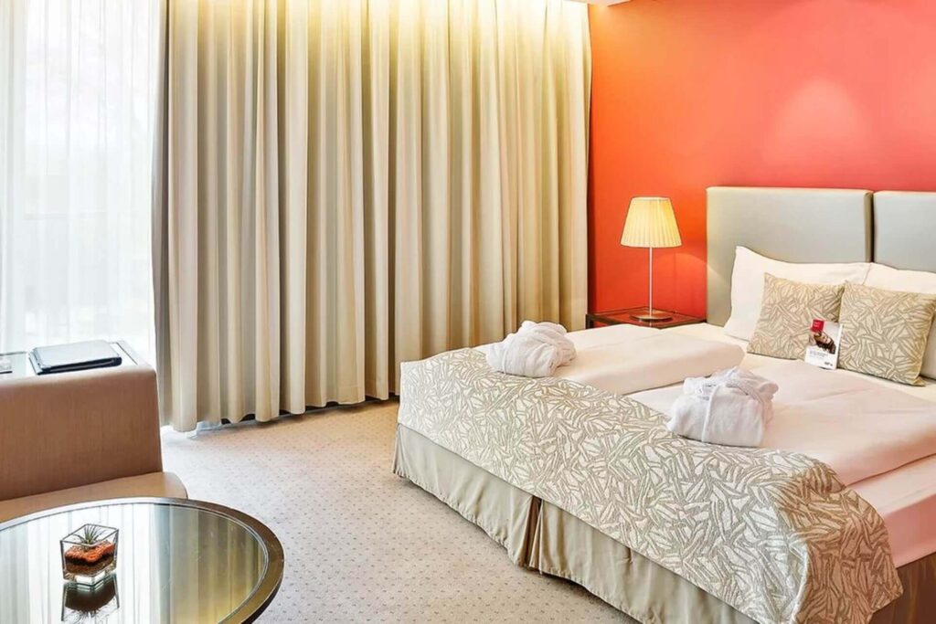 Helles Hotelzimmer mit einem Doppelbett, zwei weißen Bademänteln, gemusterter Bettwäsche, roter Akzentwand, Stehlampe, Sessel, rundem Glastisch und großem Fenster mit beigen Vorhängen - perfekt für Gäste einer Hochzeitslocation in Wien.