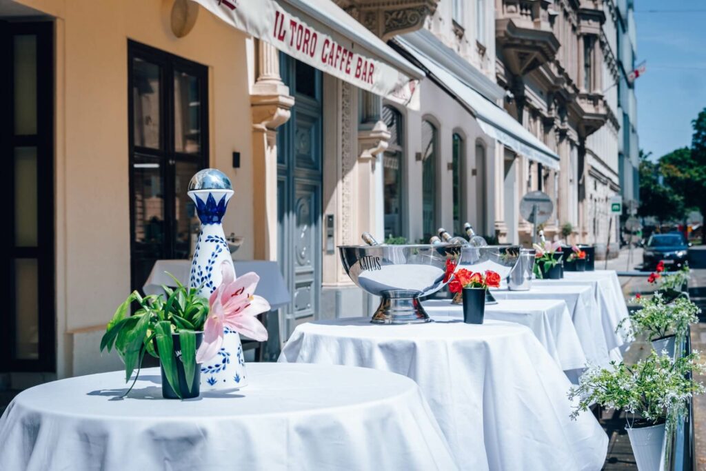Tische im Freien mit weißen Tischtüchern und Blumenschmuck bilden einen charmanten Schanigarten vor der "Il Toro Caffè Bar". Die sonnige Straße zeigt historische Gebäude und Topfpflanzen, die den einladenden Bürgersteig säumen.