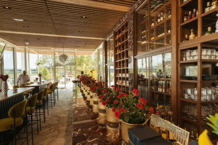 Das Interieur eines gehobenen Restaurants mit einer Reihe roter Topfpflanzen, einer Bar mit gelben Hockern, Regalen mit Glaswaren und Flaschen, Steinboden und großen Fenstern, durch die Tageslicht hereinströmt.