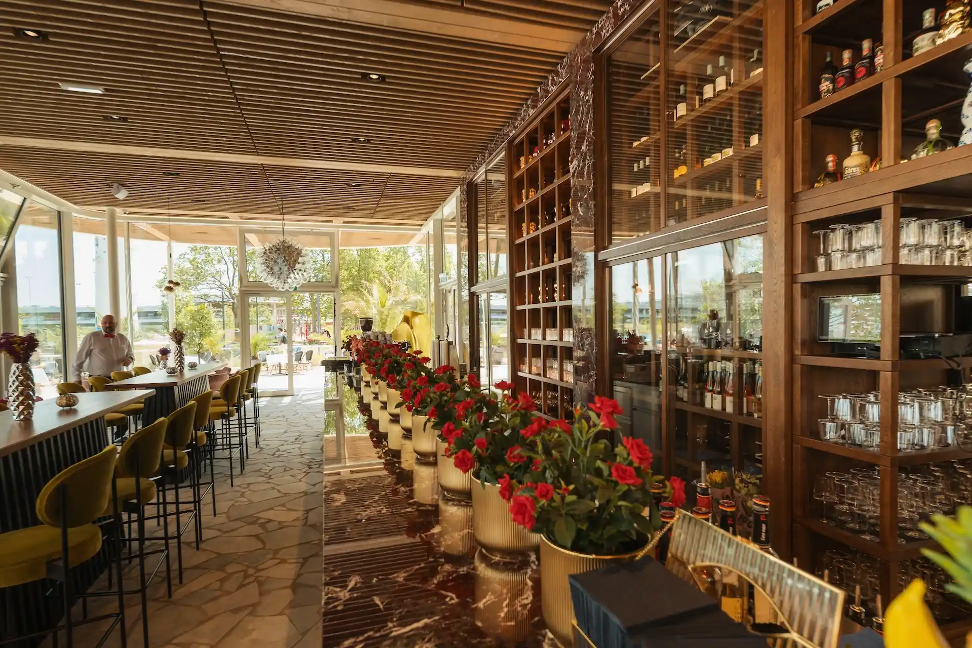 Das Interieur eines gehobenen Restaurants mit einer Reihe roter Topfpflanzen, einer Bar mit gelben Hockern, Regalen mit Glaswaren und Flaschen, Steinboden und großen Fenstern, durch die Tageslicht hereinströmt.