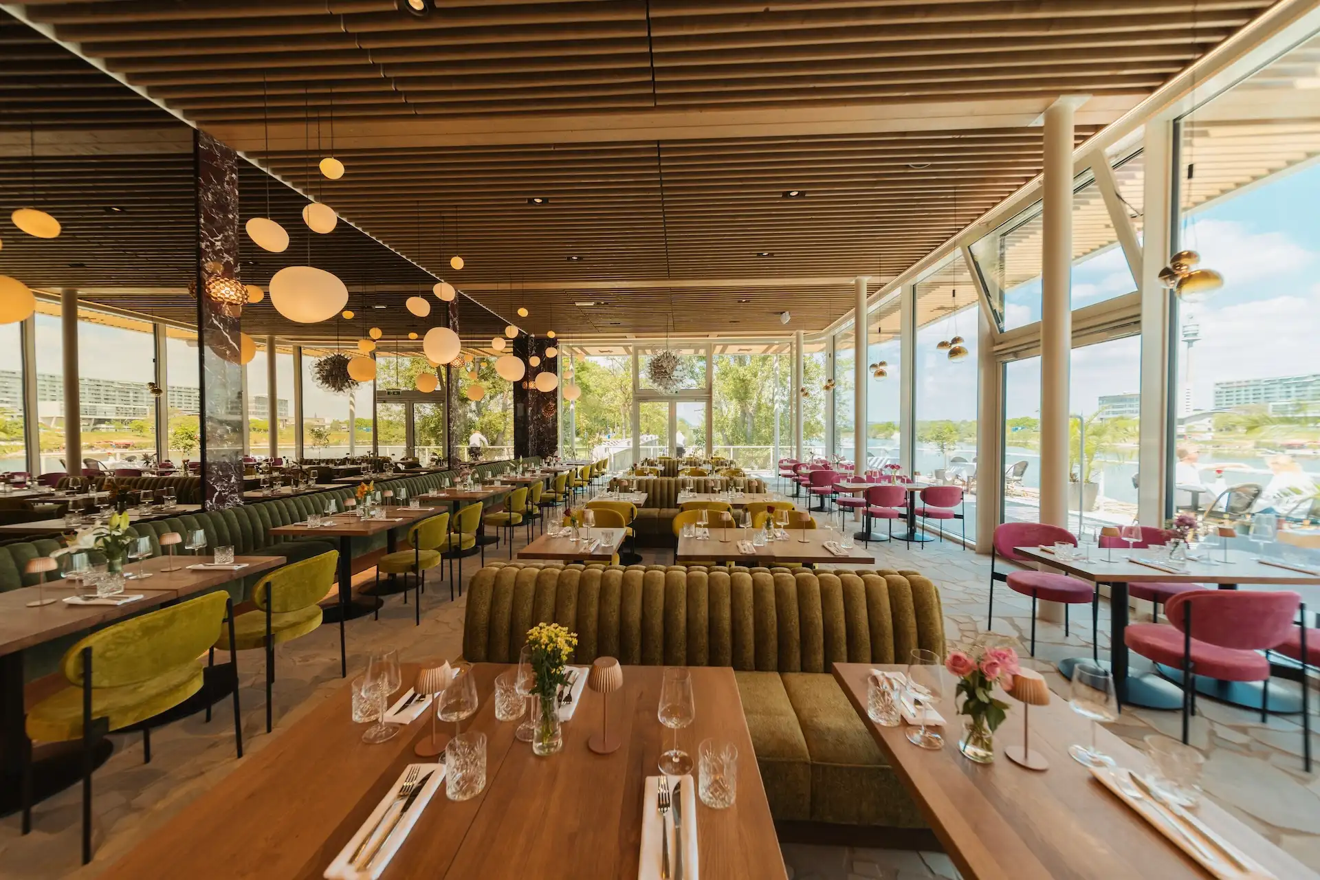 it's Amore Ein geräumiges, modernes Restaurant mit großen Fenstern, Holztischen, grünen und rosa gepolsterten Sitzgelegenheiten, hängenden Kugellampen und Blick auf Bäume und Wasser im Außenbereich. Die Tische sind mit Gläsern und Blumengestecken gedeckt.