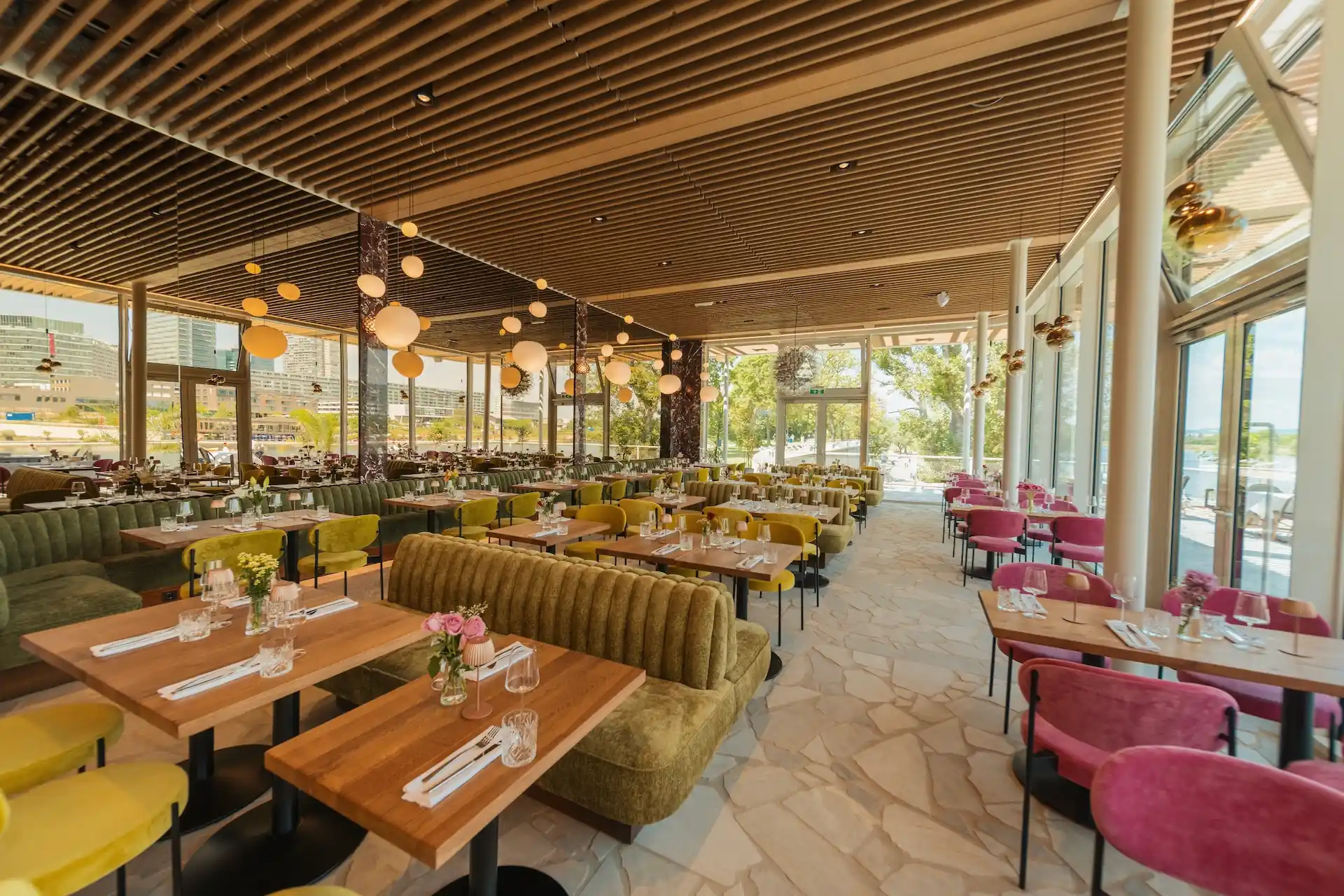 It's Amore Geräumiges, modernes Restaurant mit Holzdecke, großen Fenstern und Tageslicht. Ausgestattet mit grünen und rosa Stühlen, Holztischen, auf denen Gläser, Teller und Besteck bereitstehen, sowie hängenden runden Lampen.