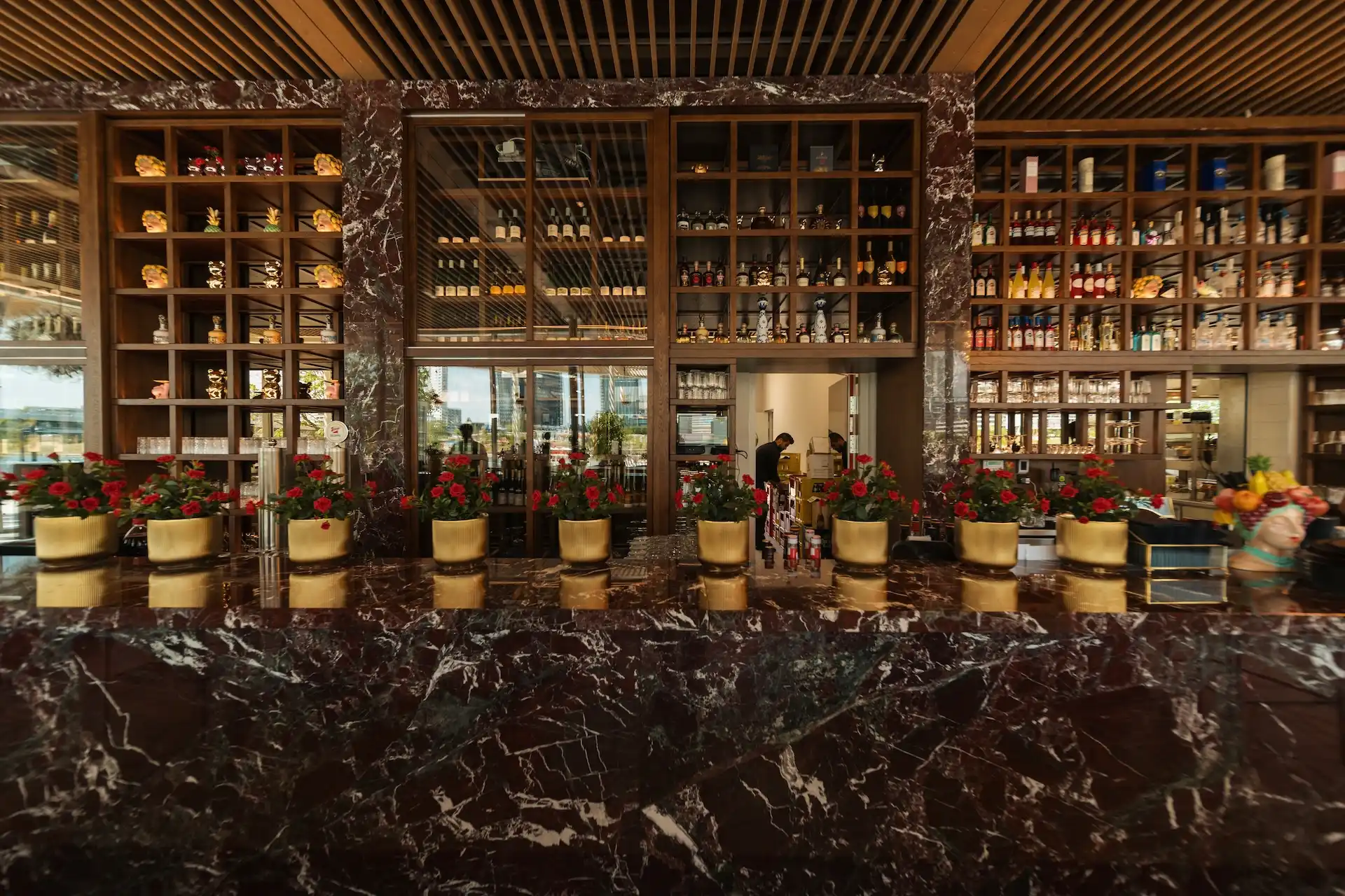 Eine Bartheke aus Marmor mit roten Topfblumen, Regale voller Flaschen und ein Spiegel, in dem sich im Hintergrund eine Person im Inneren eines stilvollen, modernen Restaurants spiegelt.