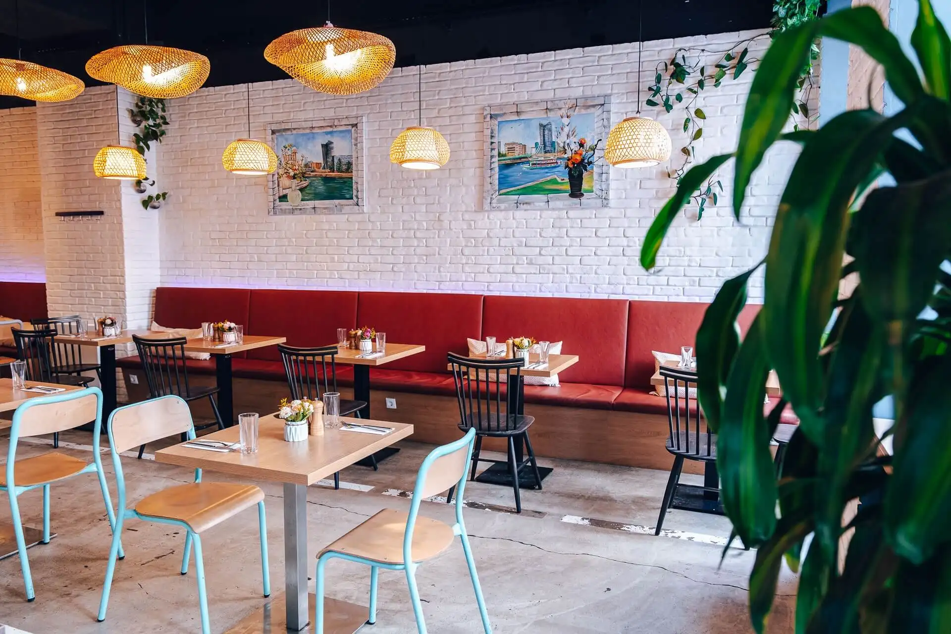 Ein modernes Restaurant mit roten gepolsterten Sitzbänken, Holztischen, schwarzen und mintgrünen Stühlen, hängenden Korblampen und gerahmter Kunst an einer weißen Backsteinwand. Im Vordergrund steht eine große Grünpflanze.