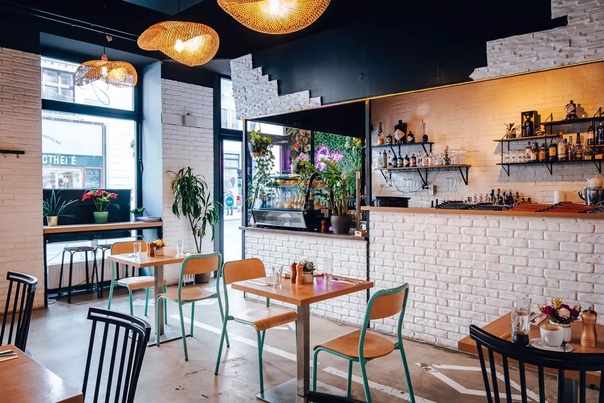 Ein modernes Café mit Holztischen und -stühlen, weißen Backsteinwänden, hängenden geflochtenen Lampen, Topfpflanzen und Regalen mit Flaschen und Gläsern hinter einem Tresen. Große Fenster lassen viel natürliches Licht herein.