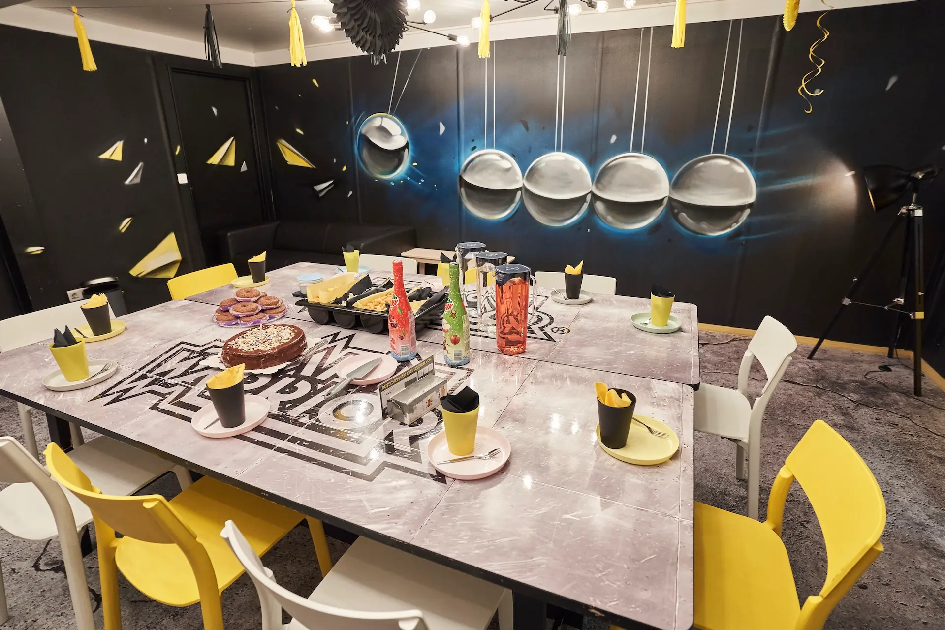 Ein für eine Party gedeckter Esstisch mit gelben und schwarzen Tassen und Tellern, Snacks und Getränken. Der Raum ist modern eingerichtet, mit einem Wandgemälde von Newtons Wiegekugeln an der Wand und gelben Akzenten überall.