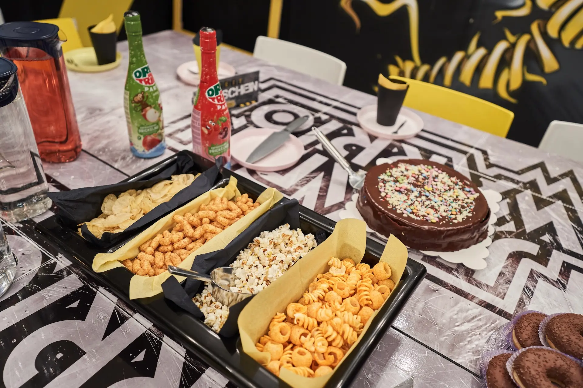 Ein Partytisch mit Tabletts mit Snacks wie Chips, Popcorn, Puffs, einem Schokoladenkuchen mit Streuseln, Cupcakes, zwei Flaschen Limonade, Tassen und Tellern auf einer gemusterten Tischdecke mit einem Dinosauriermotiv.