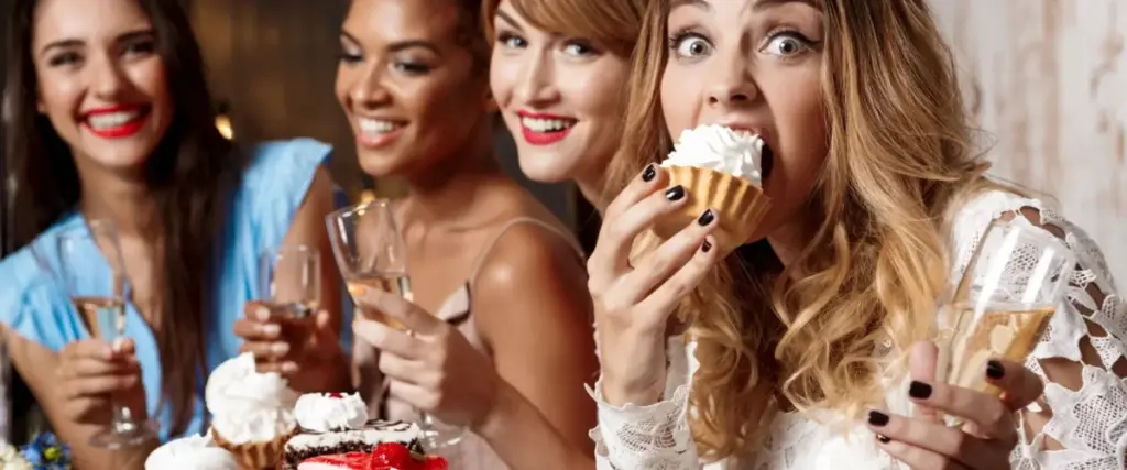 Vier gut gekleidete und lächelnde Frauen mit Sektgläsern und Desserts bei einem festlichen Junggesellenabschied in Wien. Eine Frau bereitet sich spielerisch auf den Verzehr einer mit Schlagsahne belegten Torte vor und fängt damit die fröhliche Stimmung der Feier ein.