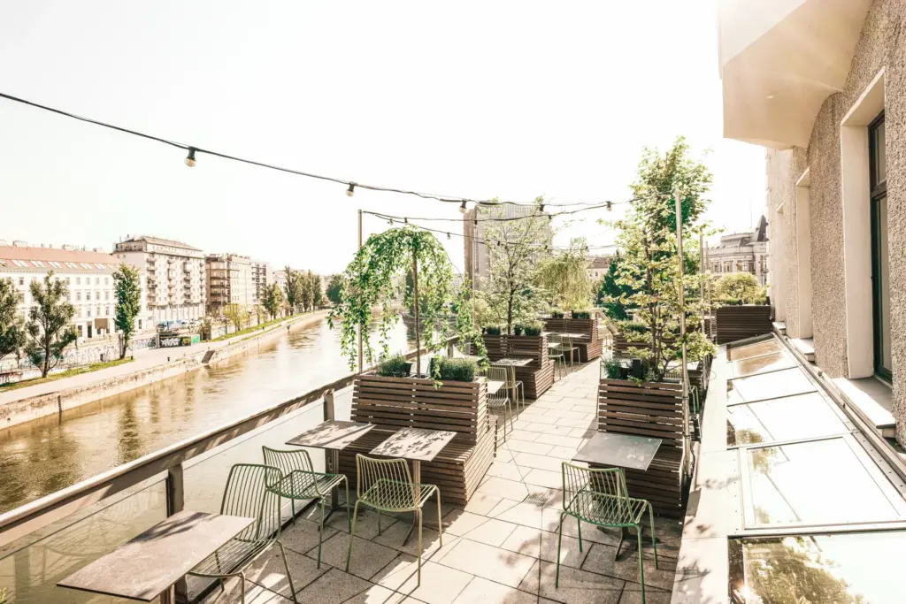 Dachterrasse mit Grünpflanzen, Holzsitzen und Metallstühlen mit Blick auf einen Fluss und städtische Gebäude an einem hellen, sonnigen Tag. Über der Terrasse sind Lichterketten aufgehängt.