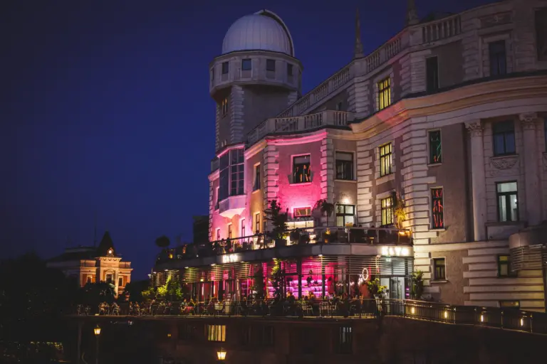 KLYO Wien - Ein großes historisches Gebäude mit einer Kuppel und einem Observatorium, das nachts mit rosafarbenen und violetten Lichtern beleuchtet wird und auf dessen Terrasse man mit Blick auf die Stadt speisen kann.
