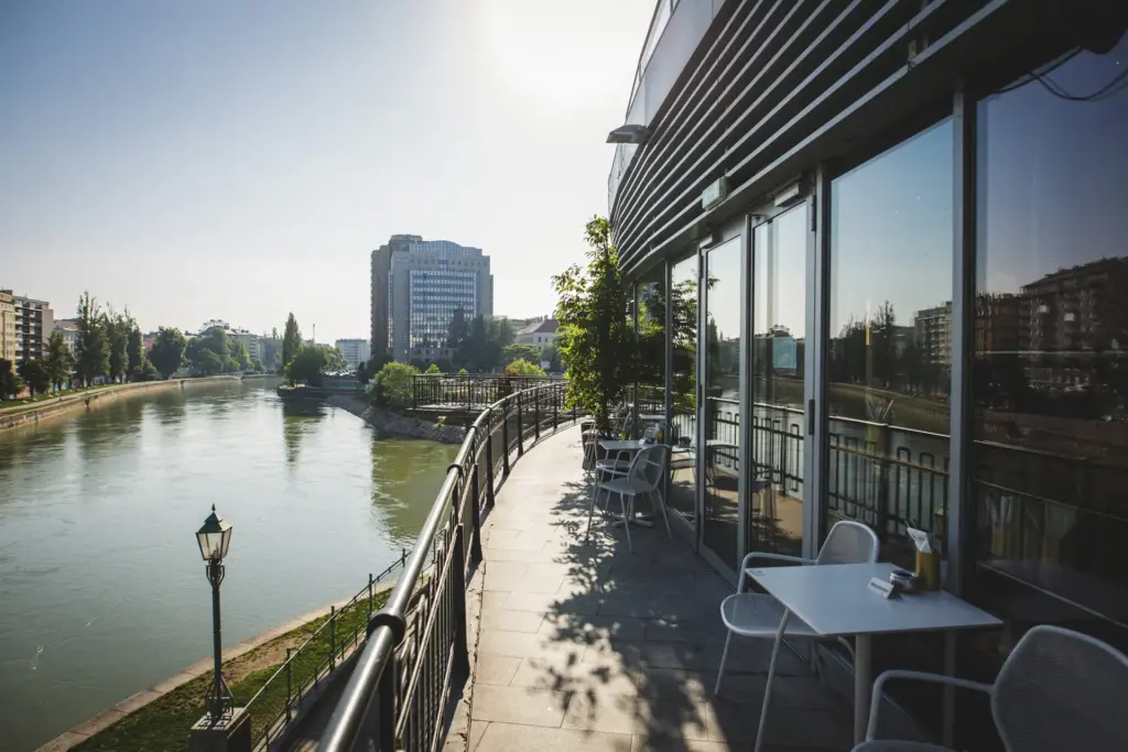 Außenterrasse mit leeren Tischen und Stühlen mit Blick auf einen ruhigen Fluss, moderne Gebäude und Bäume unter einem hellen, klaren Himmel, der sich in großen Glasfenstern spiegelt.