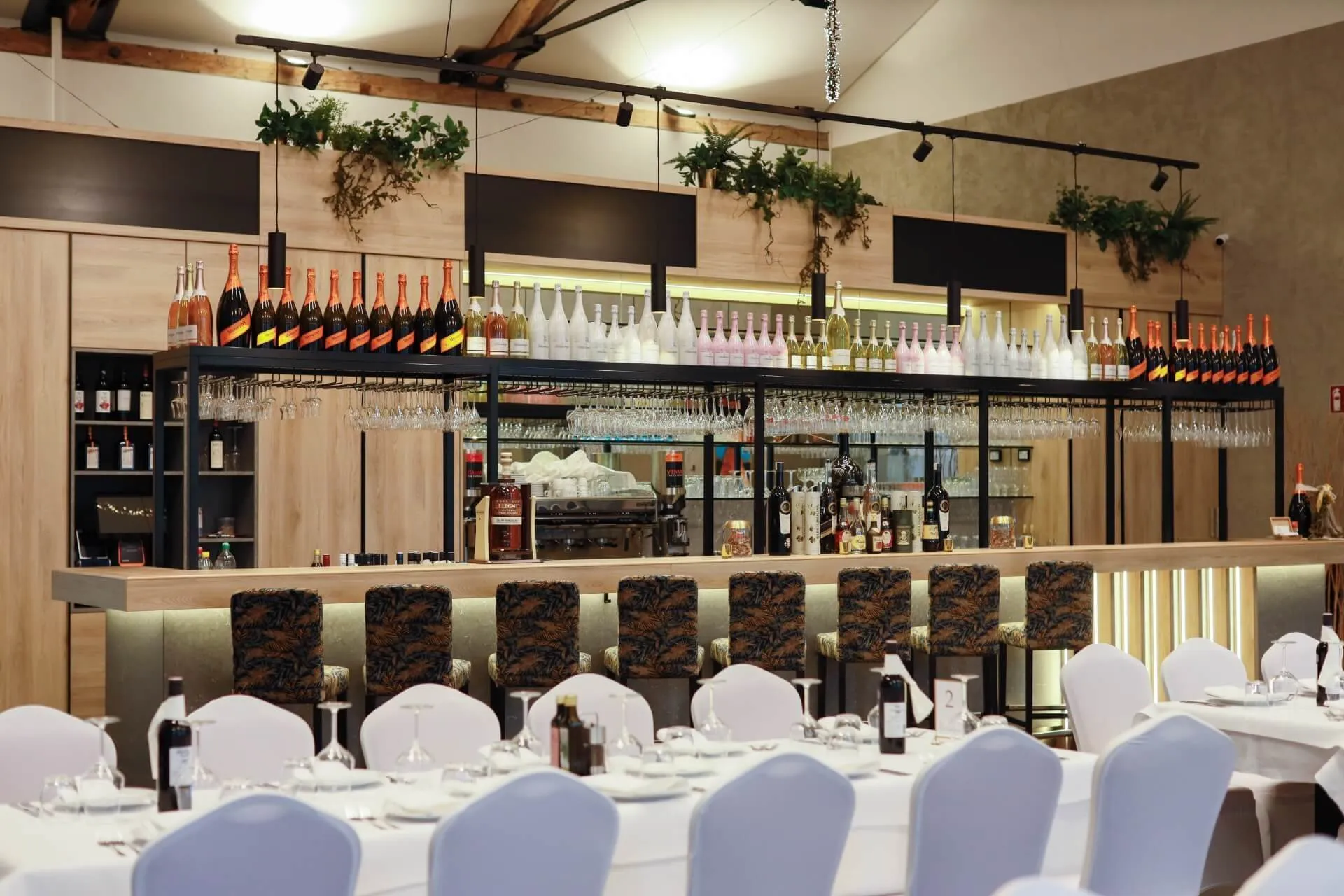 Eine moderne Bar mit hohen Stühlen, einer Reihe von Flaschen und Gläsern über dem Tresen, hängenden Weingläsern und Grünpflanzen in einem Regal. Im Vordergrund sind Tische mit weißen Tüchern und Stühlen zum Essen aufgestellt.